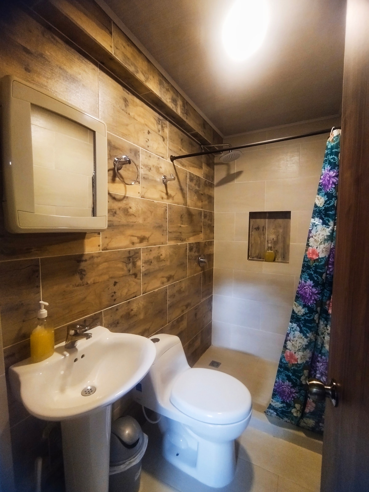 A well-maintained bathroom features a modern shower area with a colorful floral curtain. A pedestal sink and toilet are positioned against a backdrop of warm wooden tiles, contributing to a welcoming atmosphere. Ample lighting brightens the space.