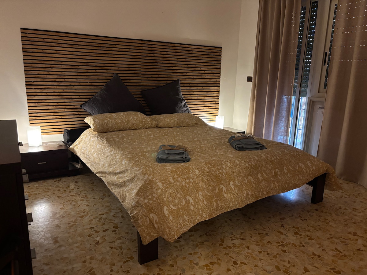 A spacious bed is positioned against a textured wooden headboard, flanked by bedside tables with softly glowing lamps. Two large pillows rest atop the bed, which is covered with a patterned duvet. Natural light filters through partially closed curtains, highlighting the warm tones of the room.