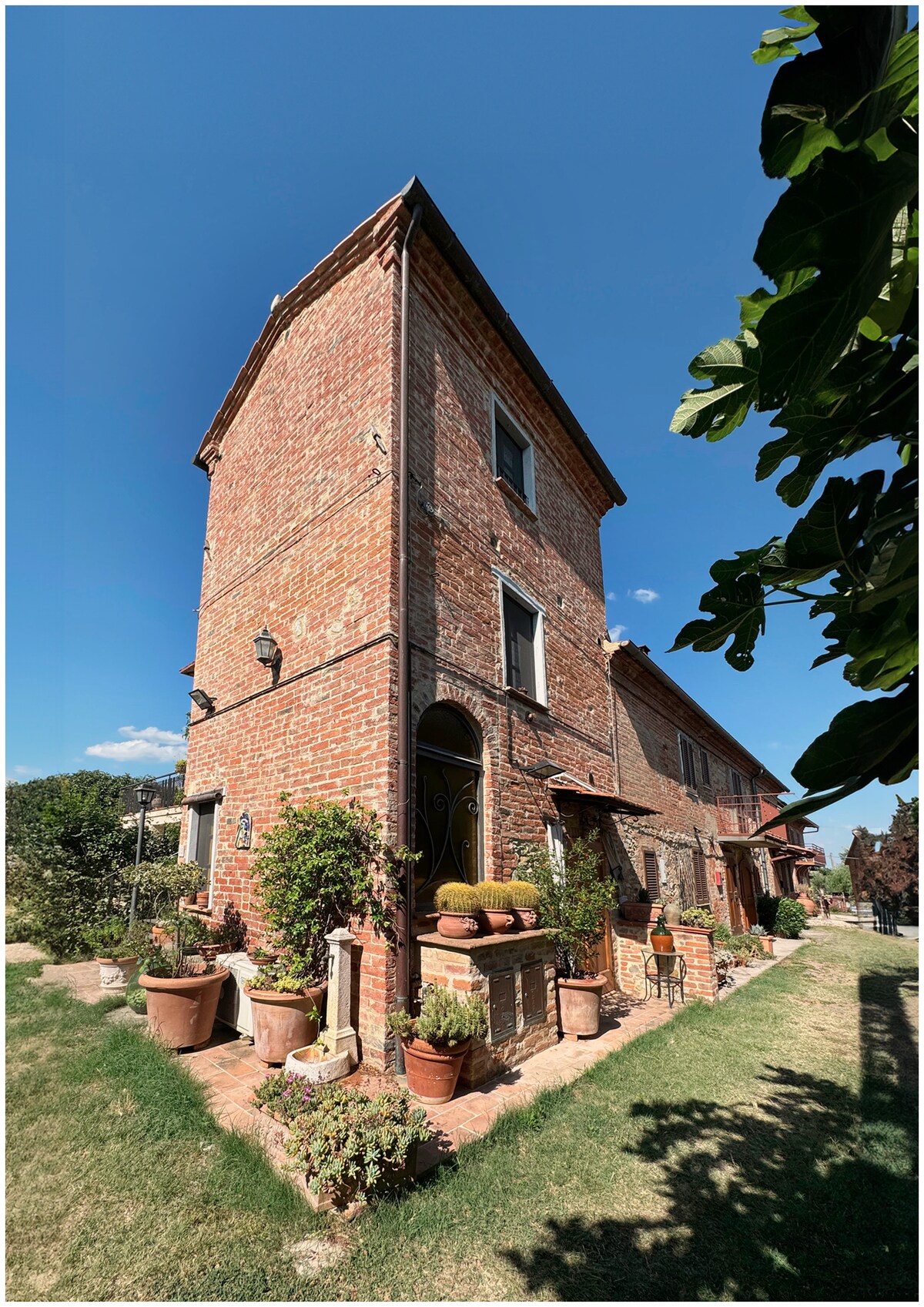 A charming brick tower stands prominently against a clear blue sky. Surrounding the base are various potted plants, showcasing greenery and flowers. The building features decorative entrance details, with lush vegetation framing the structure, and a well-maintained lawn extends beside the tower.