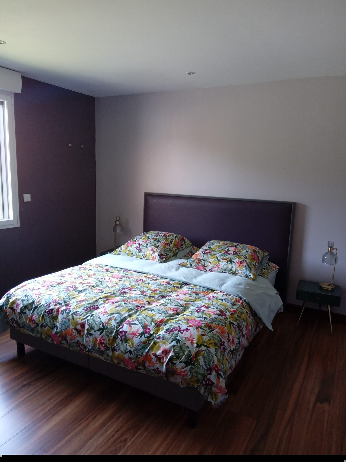 A spacious bedroom features a large bed dressed in a colorful floral duvet. Light wood flooring complements the room. A window brings in natural light, and two bedside lamps provide additional illumination. A small nightstand is positioned beside the bed.