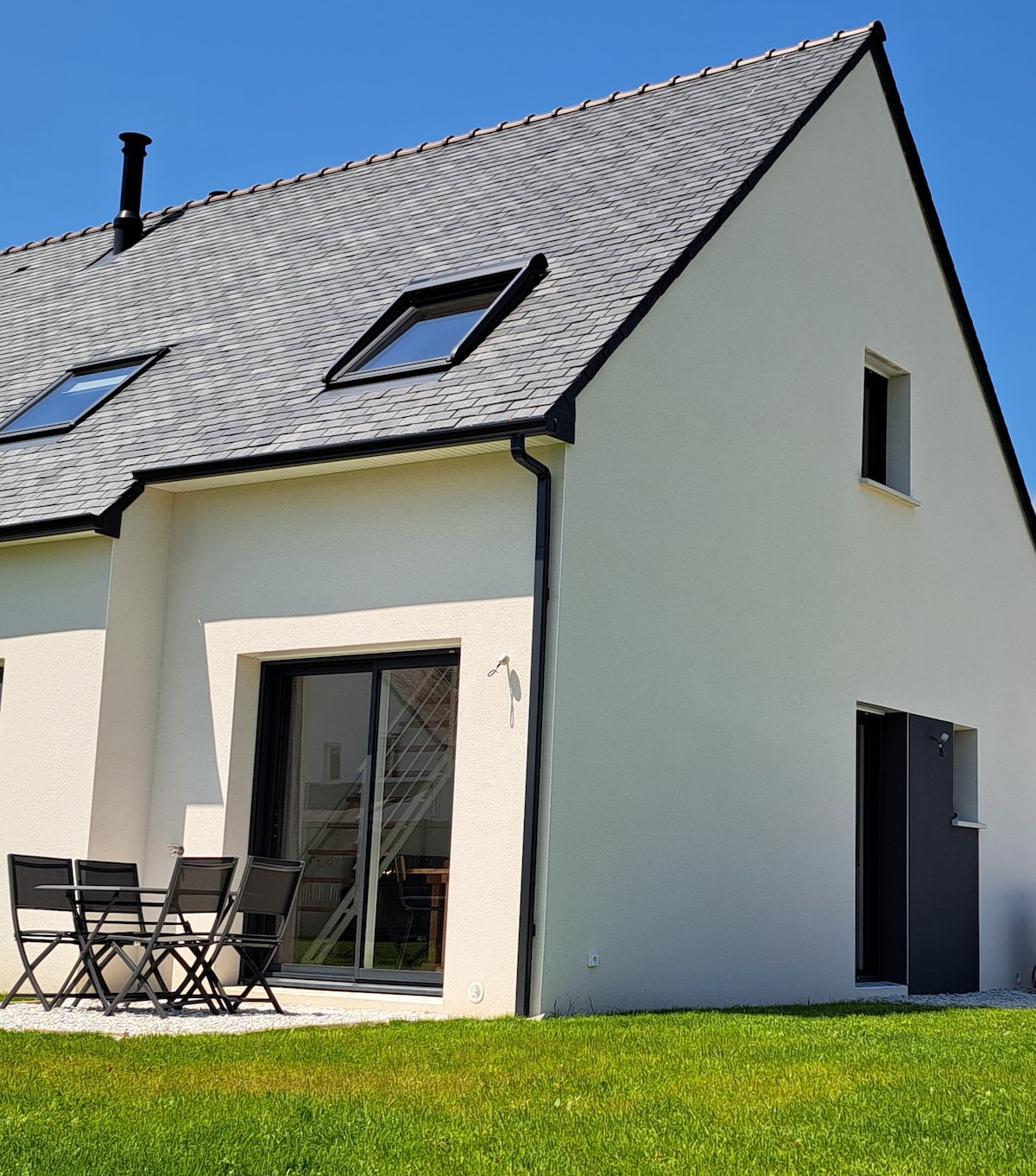 A modern house is presented from the exterior, featuring a sloped roof and large glass doors that open onto a green lawn. A small outdoor dining set with four black chairs is visible next to the entrance, creating an inviting outdoor space.