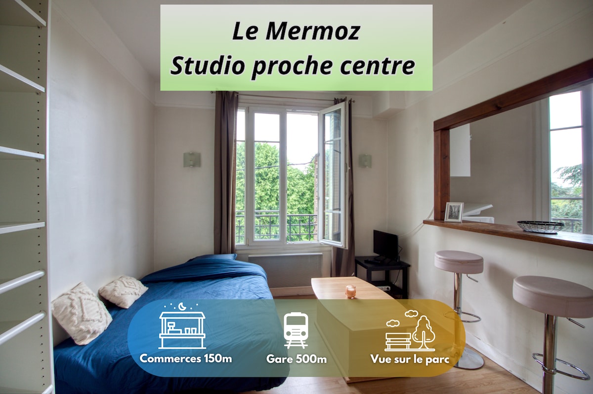 A spacious studio is shown, featuring a cozy bed with blue bedding. Natural light fills the room through large windows, offering views of the nearby park. A small table is positioned in front, with two bar stools at the separated kitchen area.