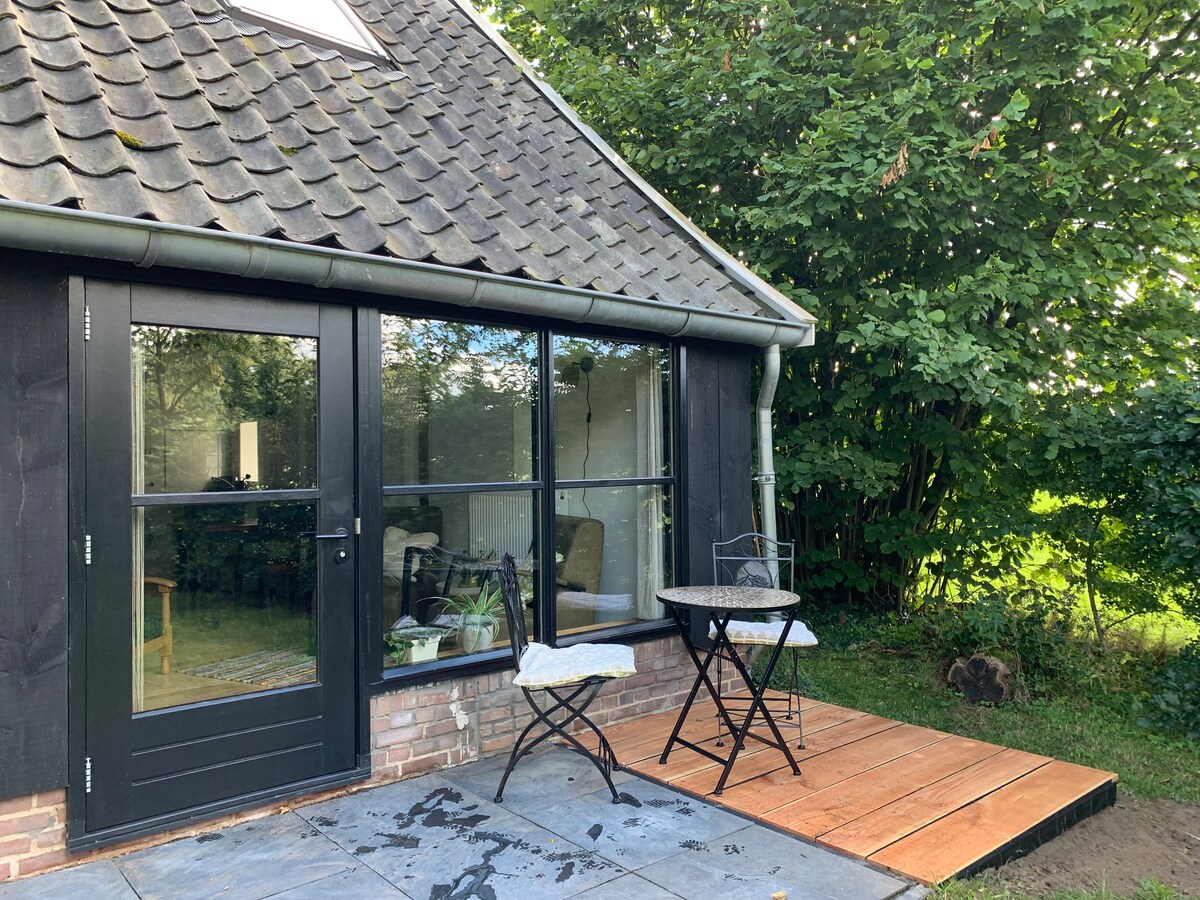 An outdoor seating area is visible featuring a small round table and two black metal chairs. The entrance to the space showcases large glass doors framed by dark wood, leading to the interior. Lush greenery borders the area, providing a sense of privacy.