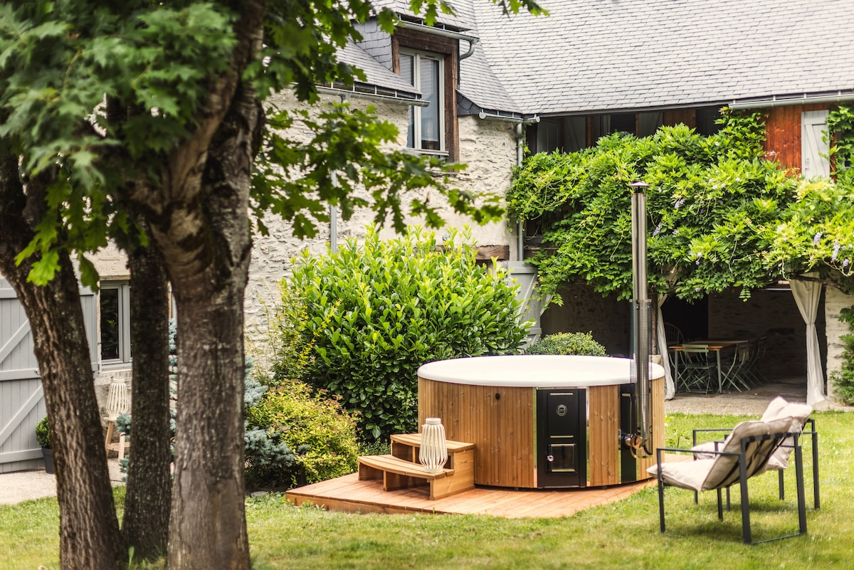 A private garden area features a wooden deck with a circular Nordic bath, surrounded by lush greenery. The bath includes a heating source, while comfortable seating is positioned nearby for relaxation.