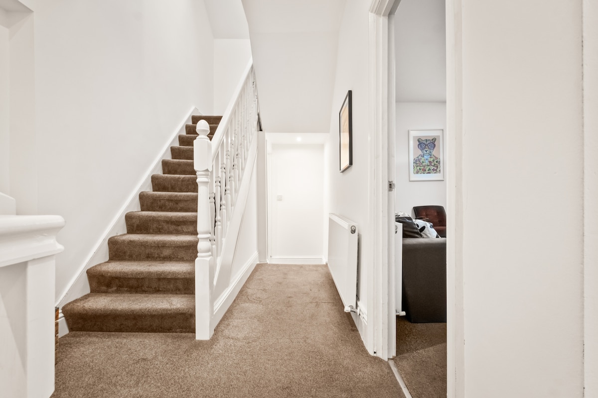 A well-lit hallway features soft beige carpeting leading up a staircase with a white banister. The bright walls are complemented by framed artwork visible in the adjacent living area. Additional doors open to various rooms, providing a sense of spaciousness and flow.