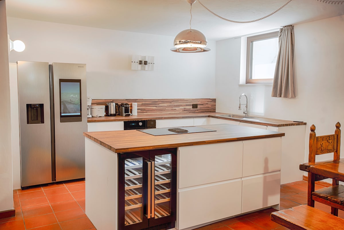 The kitchen features modern appliances, including a stainless steel refrigerator and a sleek built-in wine cooler. An ample countertop extends into a spacious island, offering a convenient cooking area. Natural light enters through a window, complemented by earthy-toned tiles and neutral cabinetry.