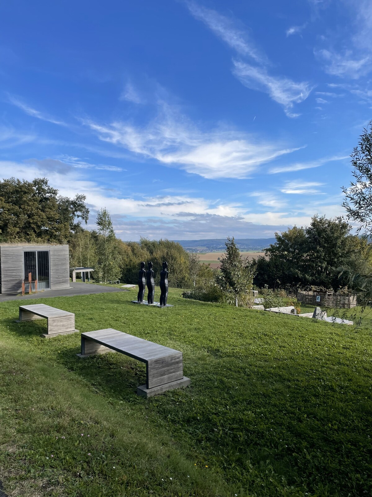 A spacious outdoor area features a manicured lawn complemented by several wooden benches. In the distance, rolling hills and trees create a natural backdrop under a blue sky with occasional clouds. A modern structure with large glass doors is visible, connecting the interior to the landscape.