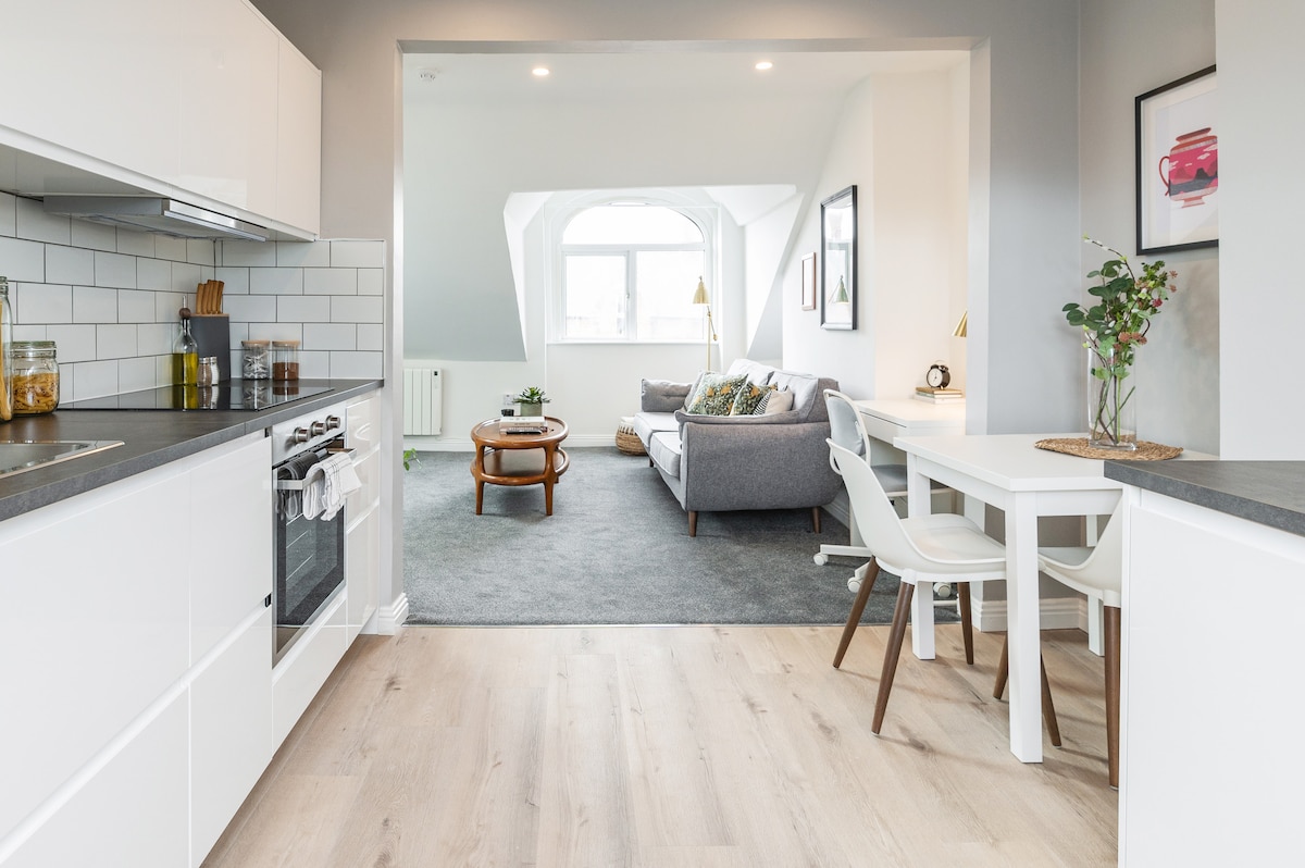 An open-plan kitchen area is visible, featuring modern cabinetry and a gas stove. A cozy living area is seen through the archway, furnished with a grey sofa, a coffee table, and natural light streaming in from large windows. A dining table and chairs are also present.