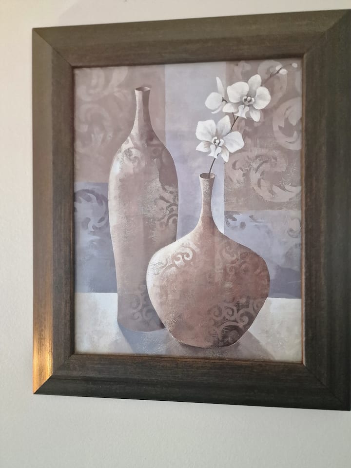 Esta elegante obra de arte enmarcada en Victoria's Classic Airbnb cuenta con dos jarrones ornamentados en tonos apagados con delicadas orquídeas blancas. El marco oscuro y resistente complementa la estética refinada, realzando el ambiente tranquilo y elegante de la habitación.