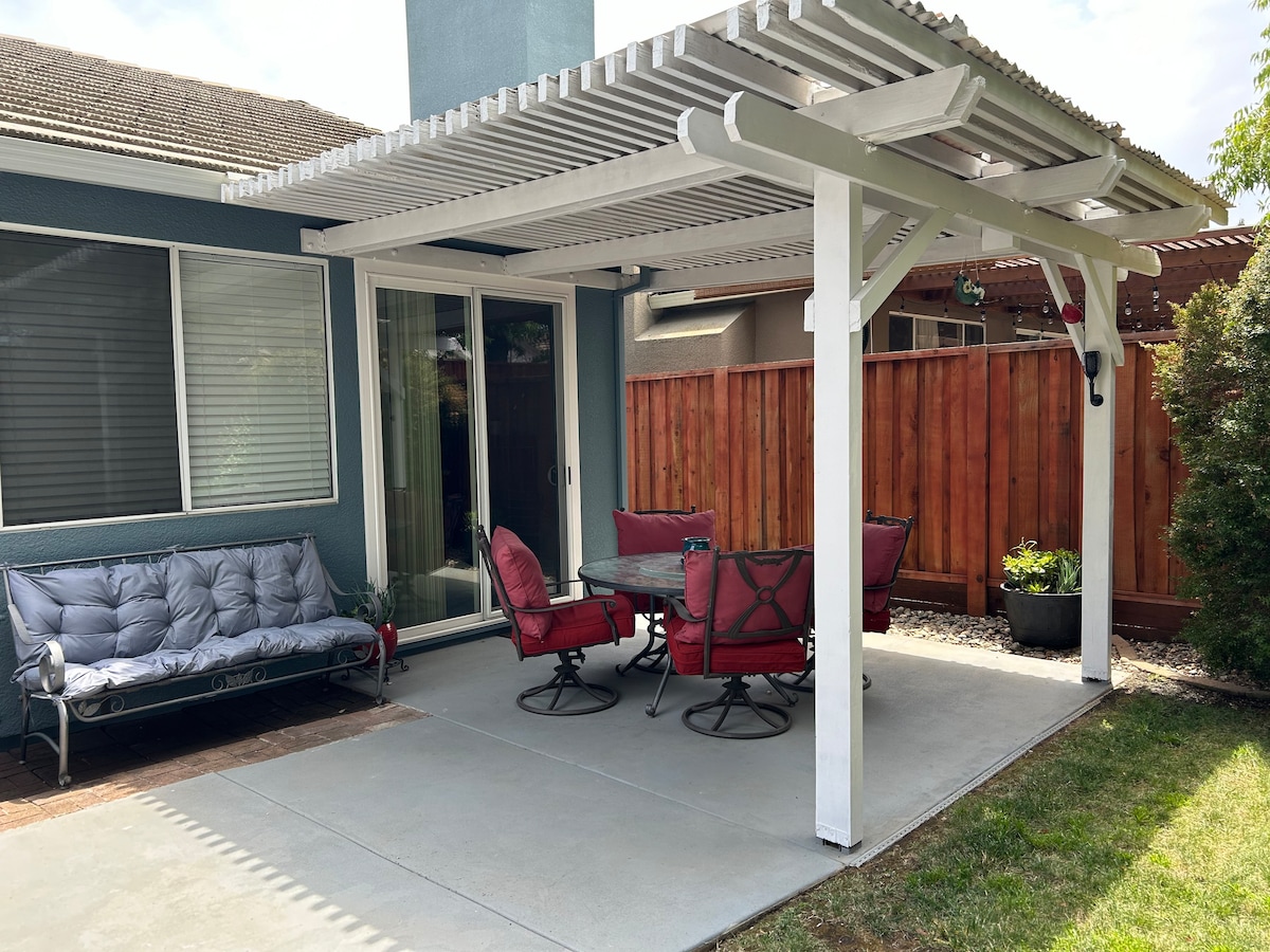 A shaded outdoor patio area features a comfortable seating arrangement with a sofa and four dining chairs around a circular table. Lush greenery borders the space, and the fence provides privacy, while soft light filters through the pergola above.