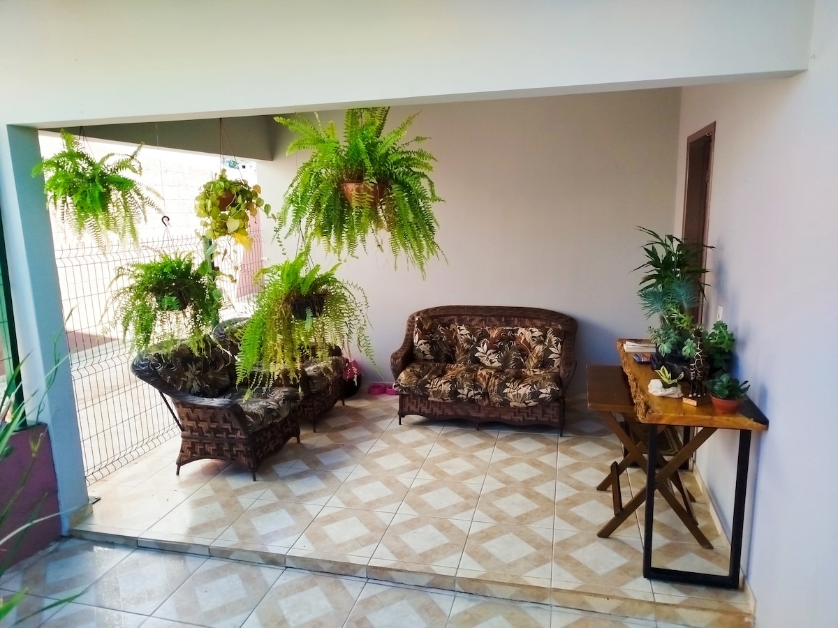 A welcoming outdoor space features two patterned sofas complemented by hanging potted ferns. A wooden table is positioned nearby, completing the tranquil atmosphere. The tiled floor enhances the area, which is well-lit by natural light filtering through the open entryway.