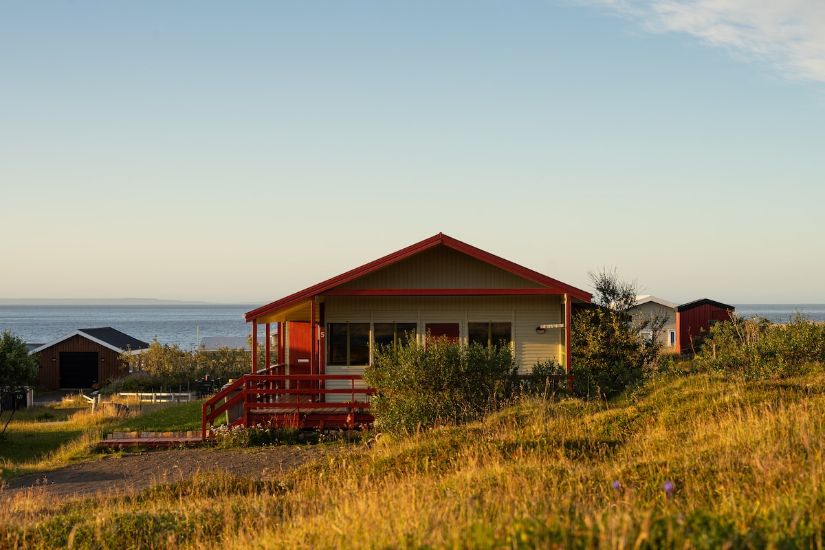A simple cabin is set against a backdrop of the sea, featuring a red-roofed porch and a pathway leading to the entrance. Surrounding greenery adds a touch of nature, while the horizon displays a soft sky, suggesting tranquility in the enchanting village of Bakkafjörður.