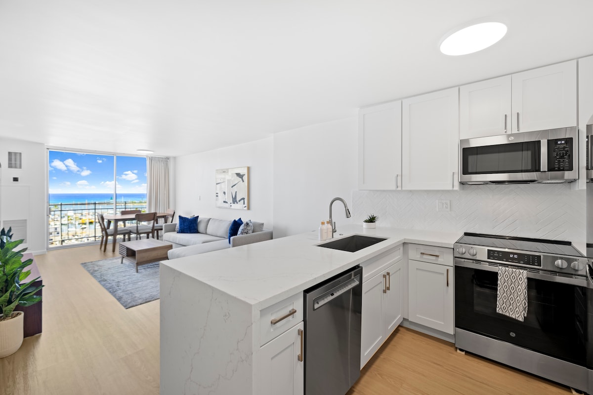 An open-concept kitchen and living area are showcased, featuring modern appliances and a spacious island. Natural light brightens the room through large windows, revealing ocean views. A comfortable sectional sofa and dining area can also be seen, creating a welcoming space for relaxation.