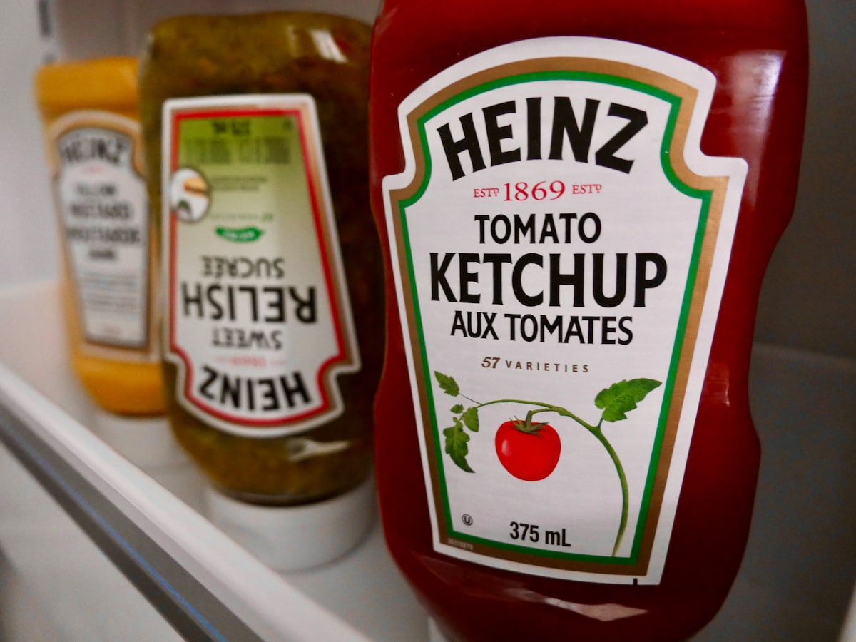 Various condiment bottles are arranged on a refrigerator shelf, prominently featuring a bottle of Heinz Tomato Ketchup in the foreground. The ketchup label displays the brand name and details, while other bottles of sweet relish and mustard are slightly blurred in the background.