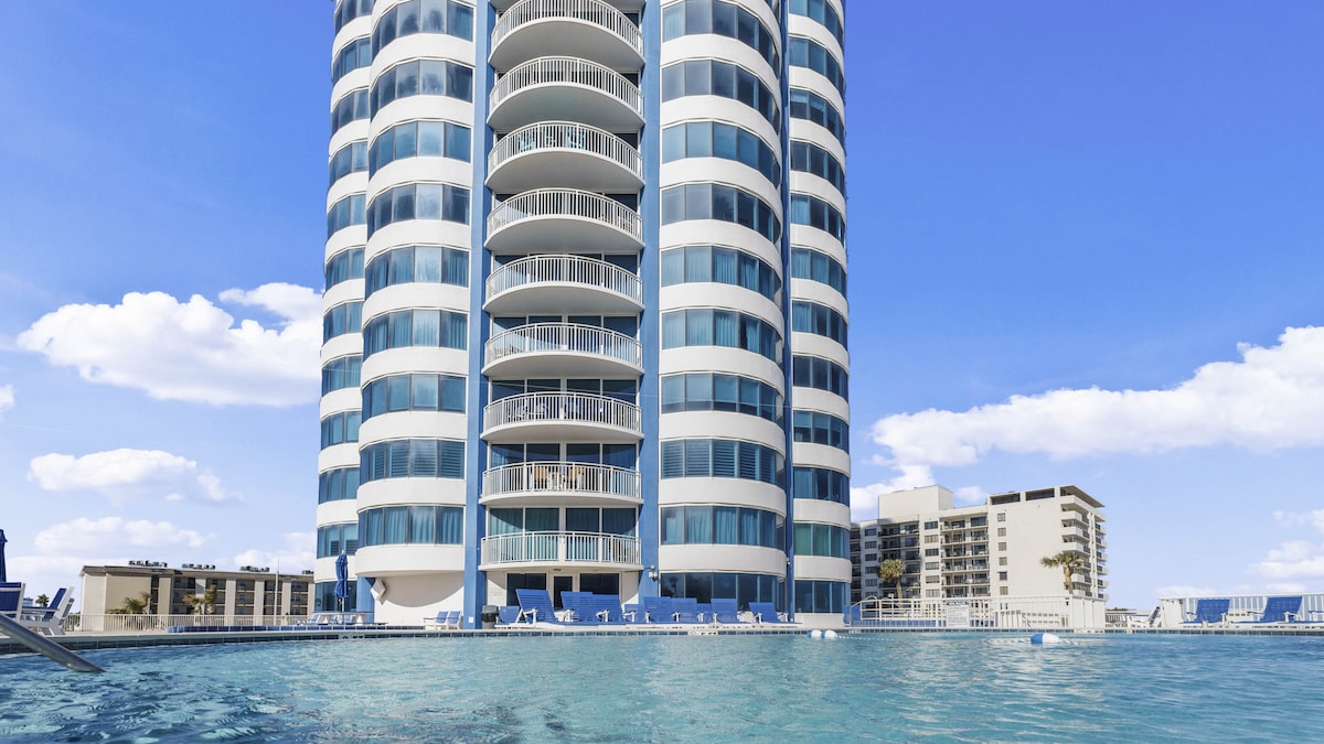 A modern high-rise building features a curved design with numerous glass balconies. A clear pool reflects the structure, surrounded by lounge chairs. The backdrop shows a clear blue sky with a few clouds, enhancing the open feel of the outdoor space.