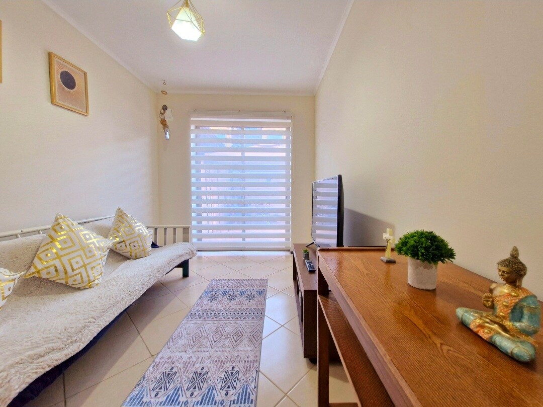 A bright living area with white walls features a cozy couch adorned with decorative cushions. A wooden table stands nearby, accompanied by a small potted plant. A flat-screen TV is prominently displayed, and light filters through the elegant blinds, creating a warm ambiance.