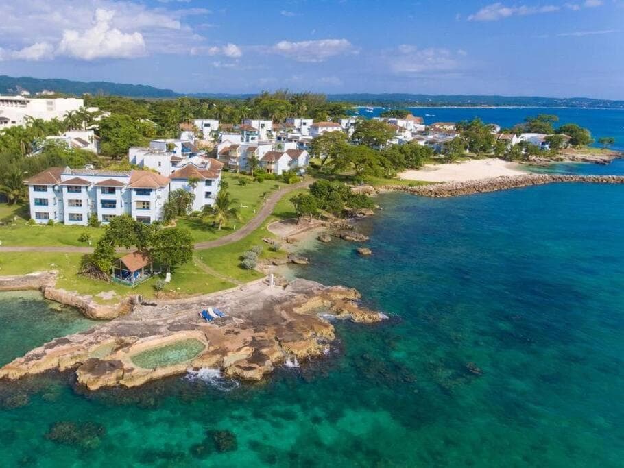 Aerial view showcases a vibrant coastal landscape featuring a series of white structures nestled among green trees. The clear turquoise waters surround rocky shoreline, leading to a sandy beach, all under a bright blue sky with scattered clouds.
