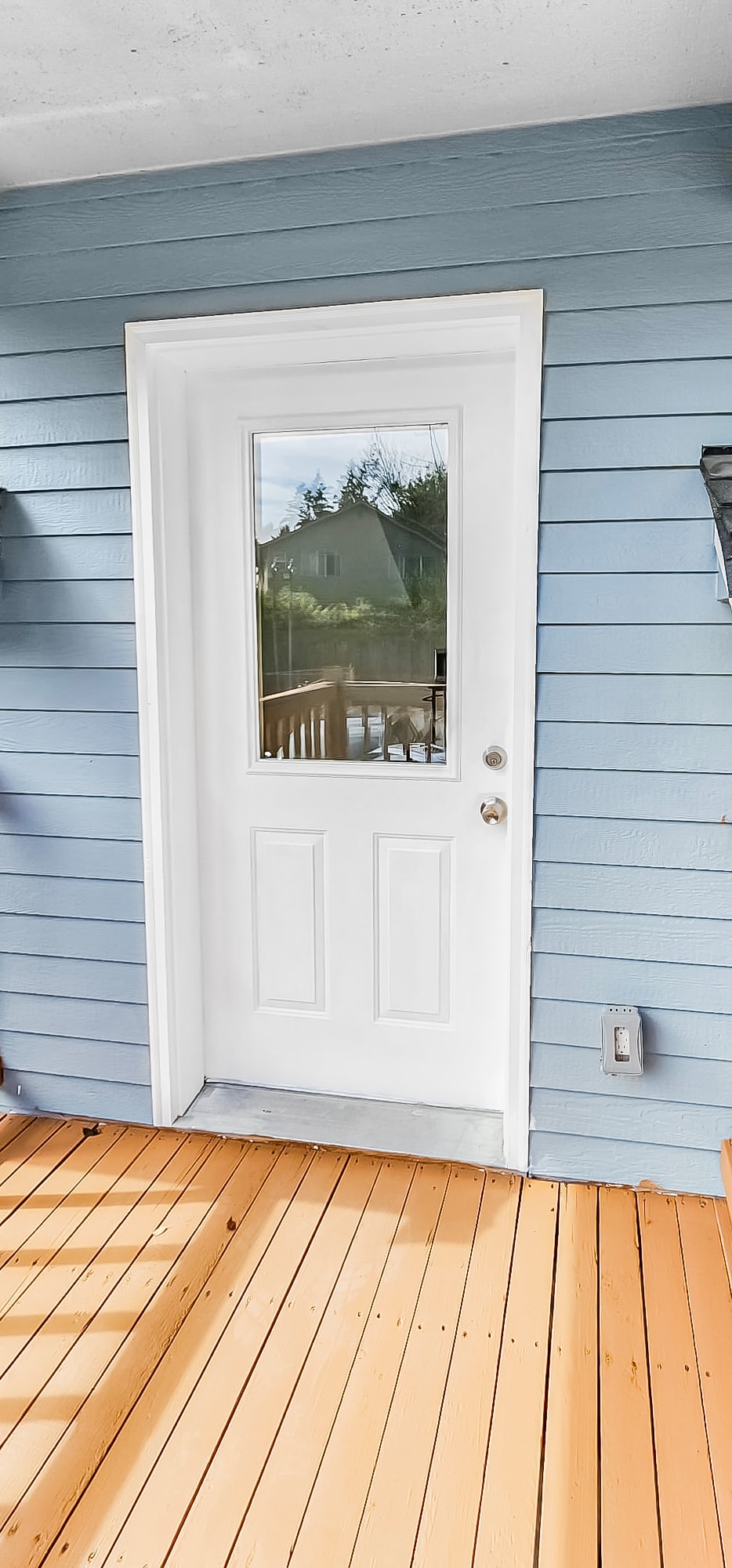 A white door with a glass panel is set against a light blue wooden wall. Natural wood decking leads up to the entrance, reflecting soft light. A wall-mounted light fixture is positioned to the right of the door, contributing to the inviting exterior.