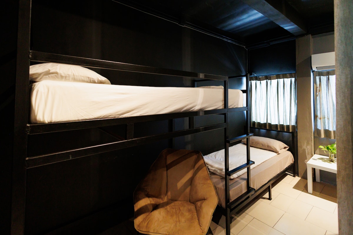 A cozy room features a bunk bed with a spacious bottom sleeping area. A large beanbag chair is positioned in the corner, and natural light filters through two windows dressed in sheer curtains. The dark walls and ceiling create a modern and intimate ambiance.