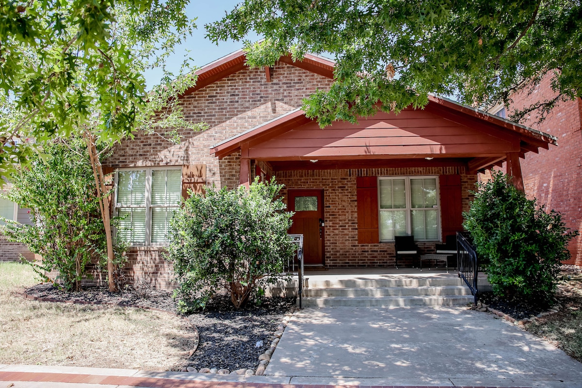 The exterior of the home exhibits a welcoming brick facade with a covered porch and red accents. Two mature trees provide shade in the front yard, while bushes frame the entrance. A concrete walkway leads to the front door, enhancing accessibility.