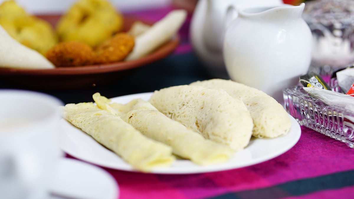 A plate is set with rolled pancakes, showcasing a light and fluffy texture. Surrounding items include various local dishes, all presented on a vibrant woven tablecloth. The setting suggests a warm, communal dining experience, perfect for enjoying a daily breakfast.