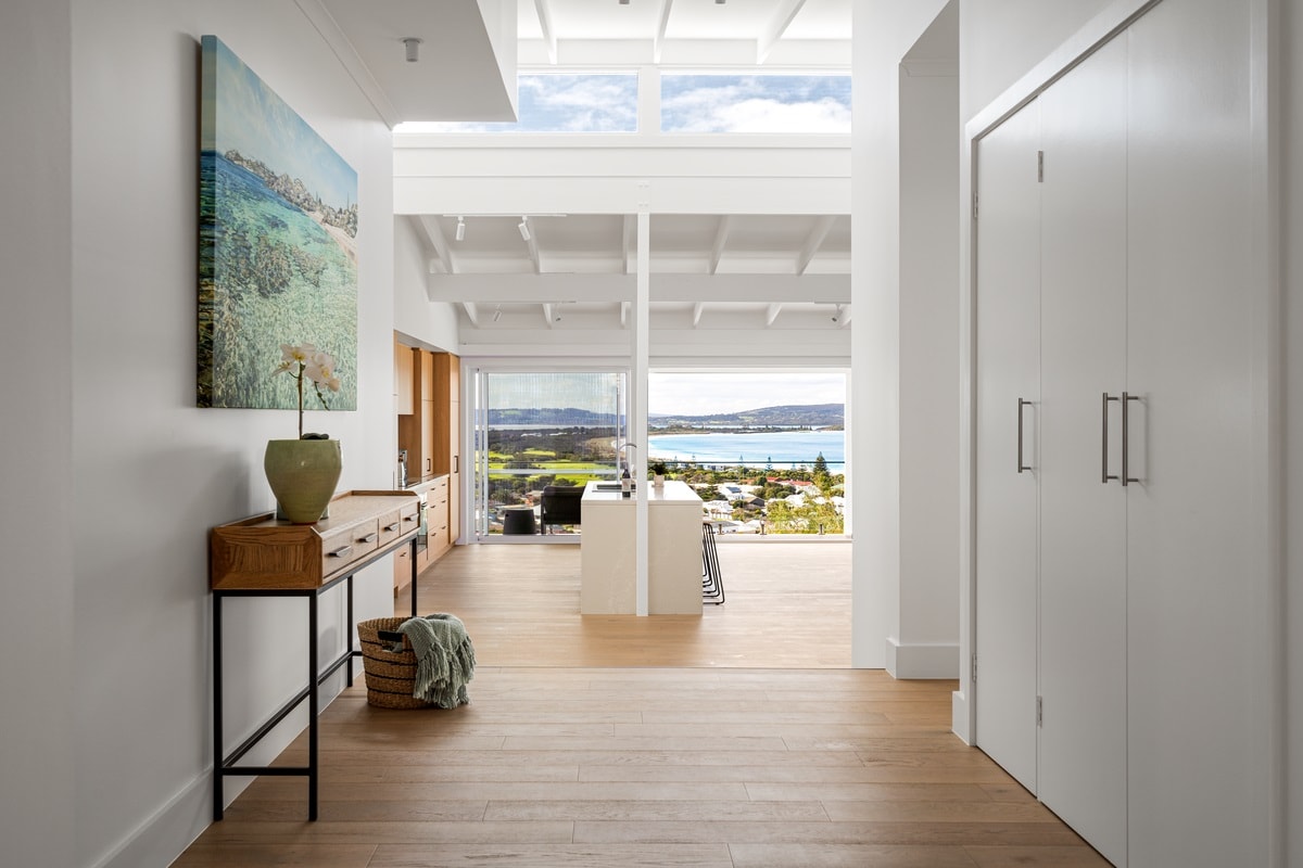An airy hallway is presented with light-colored wooden flooring and high ceilings. A large painting of a coastal scene is displayed on the wall, complemented by a wooden console table with a decorative vase. Views of the coastline can be seen through expansive windows.