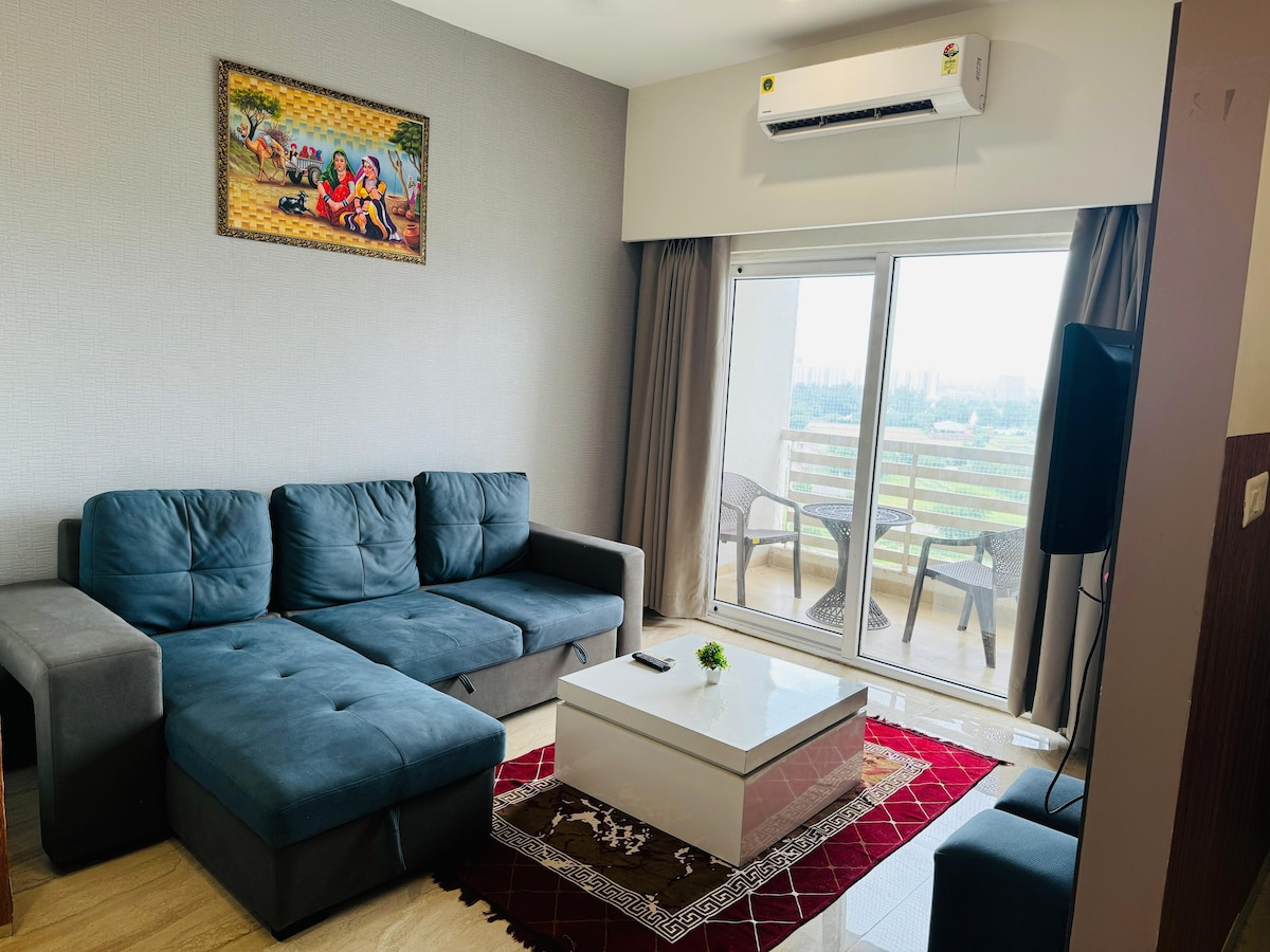 A modern living area is showcased, featuring a comfortable sectional sofa in shades of blue. A glass coffee table is placed on a decorative rug, while large sliding doors lead to a balcony with outdoor seating. An air conditioning unit is mounted on the wall.