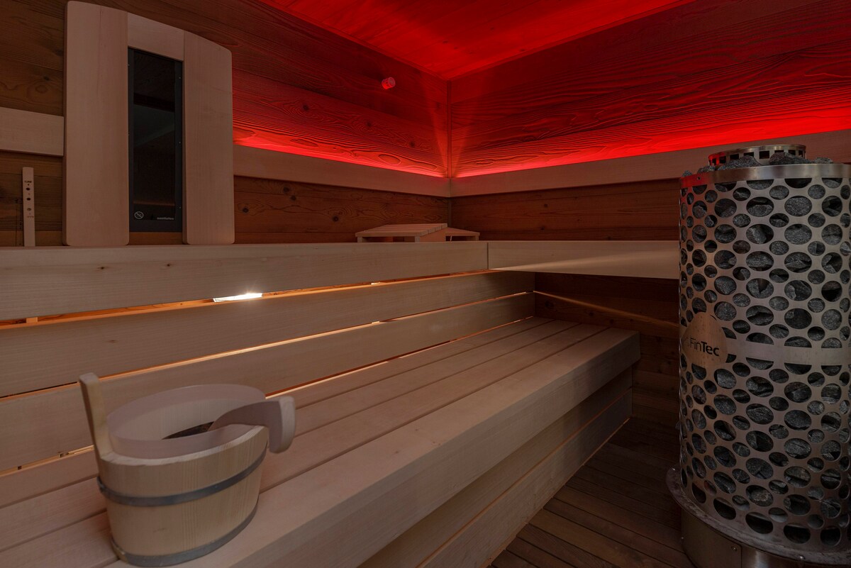 The sauna features light wooden panels and benches, offering a warm and inviting space. Soft red lighting enhances the ambiance. A metal heater stands against the wall, providing warmth to the area. Simple wooden accessories such as a bucket and ladle are present.