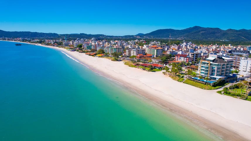A panoramic view of Canasvieiras beach showcases soft, sandy shores lined by residential buildings and local establishments. The calm, turquoise waters gently lap against the beach, framed by distant mountains under a clear blue sky.