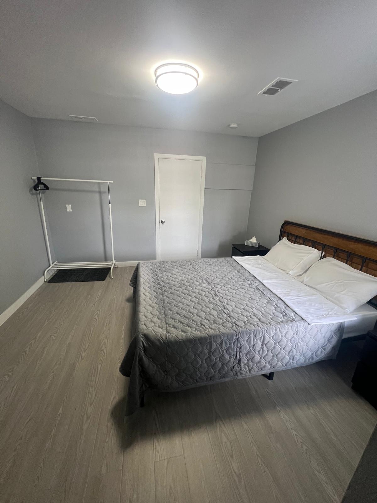 A neutral-toned bedroom showcases a spacious bed with a gray quilt. Two white pillows are neatly arranged on the bed, while a simple black nightstand is positioned beside it. A clothing rack stands against the wall, and a door leads to another area.