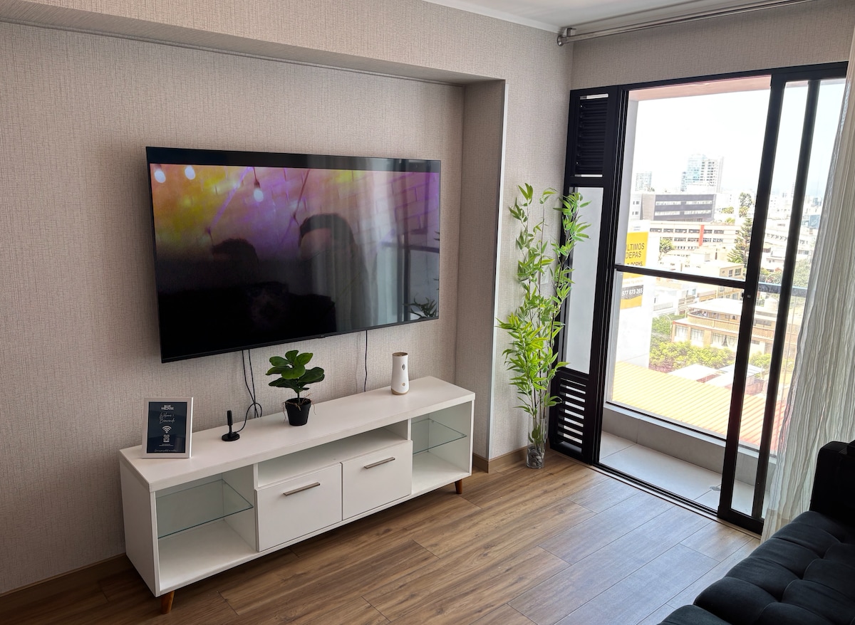 A living area features a large wall-mounted television displaying vibrant visuals. Below, a sleek white media console provides storage, accompanied by a small indoor plant for a touch of greenery. Natural light filters through a window, revealing an urban view.