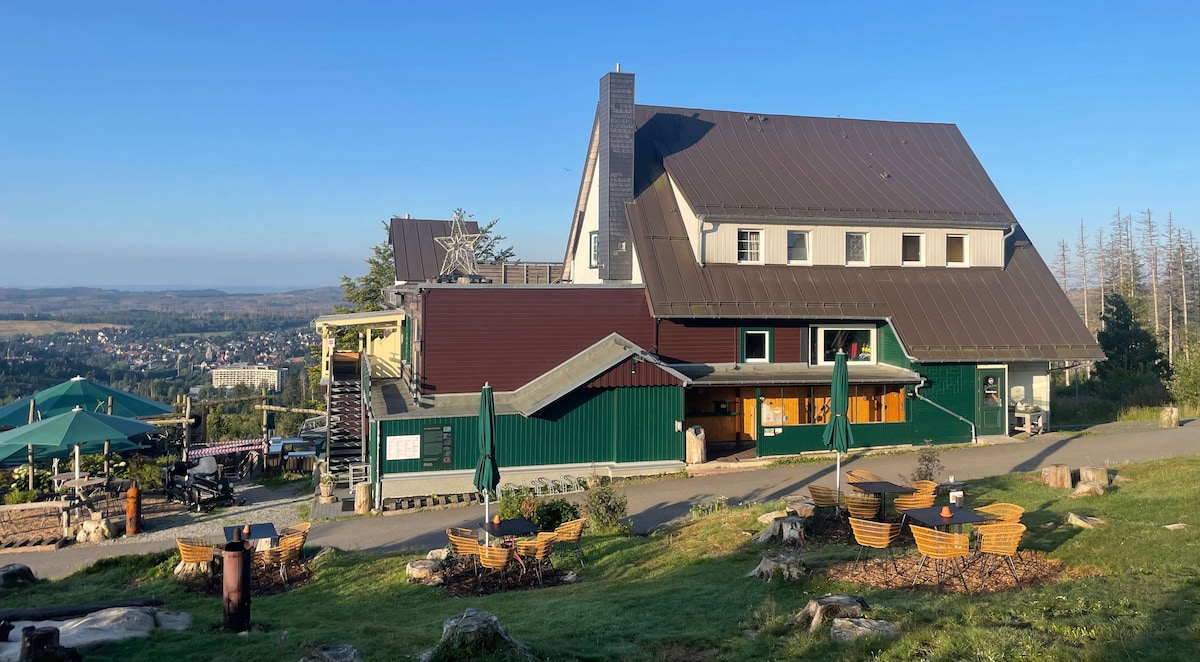 The exterior of the building is shown with a sloped roof and large windows, nestled amidst trees and open space. Outdoor seating areas with umbrellas are visible, along with scenic views of the landscape in the background under a clear blue sky.