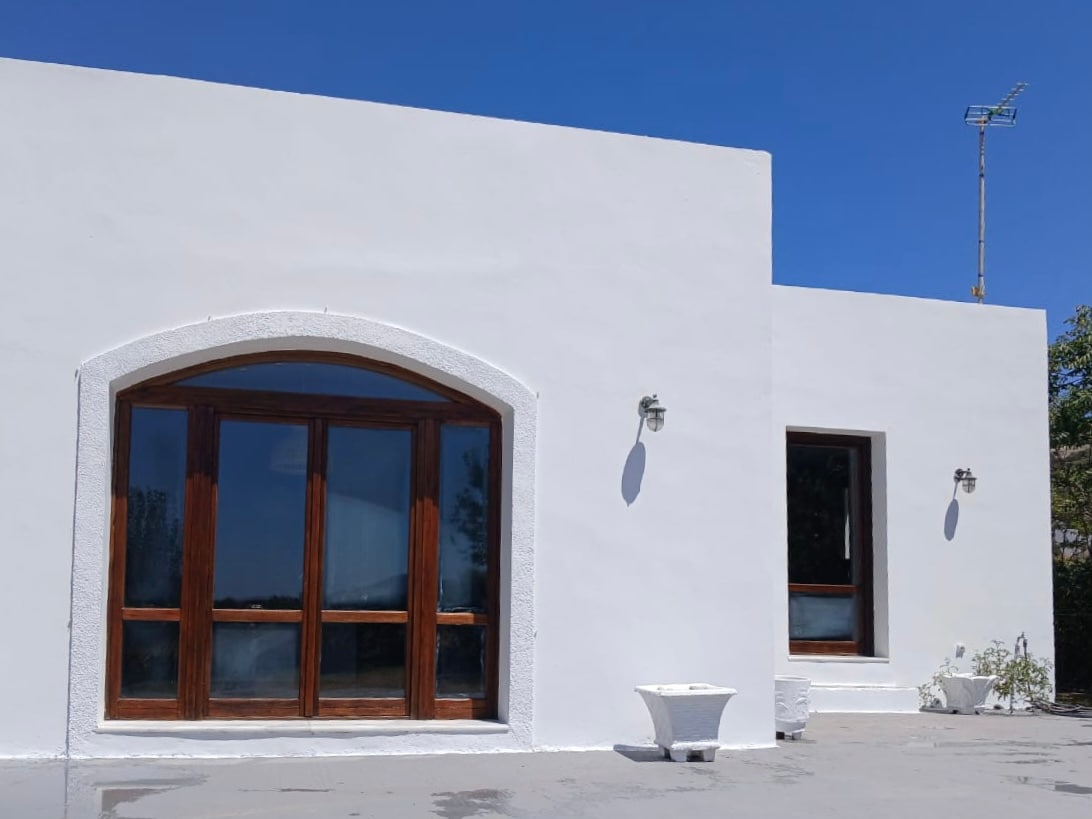 The exterior of the residence features smooth white walls and large wooden framed windows that allow natural light to illuminate the interior. A small white table sits on the patio area, providing a space to enjoy the surrounding views.