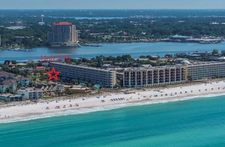 An aerial view captures a beachfront complex with a distinctive star marking Unit 215. The scene showcases a sandy beach leading to clear turquoise waters, framed by coastal buildings and distant greenery along the shoreline, providing insight into the unit's proximity to the beach.