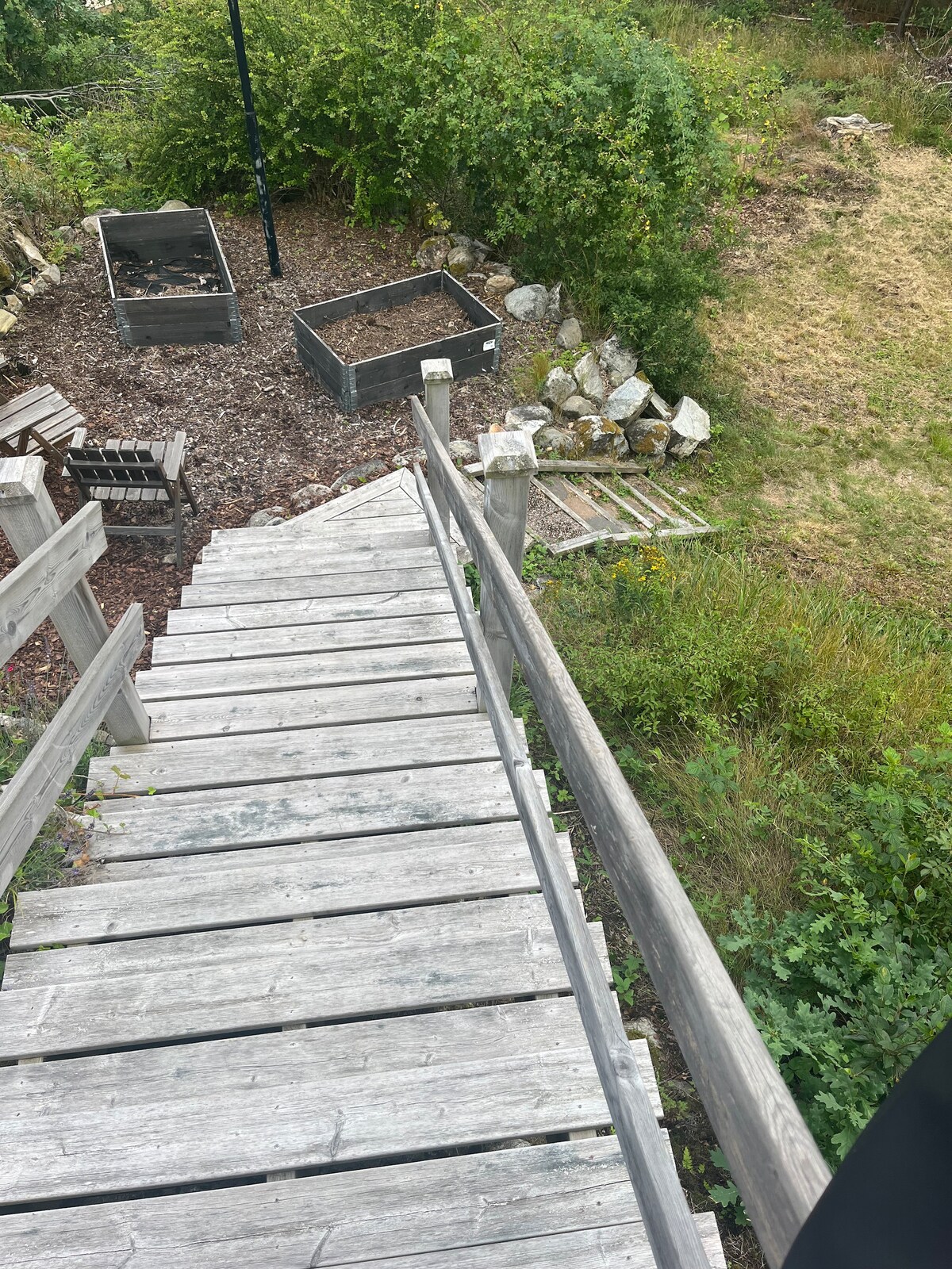 A wooden staircase leads down through a natural setting, surrounded by greenery and manicured garden beds. To the side, rocky elements and mulch contribute to a rustic atmosphere. The surrounding area features uncut grass and foliage, adding to the tranquil outdoor environment.