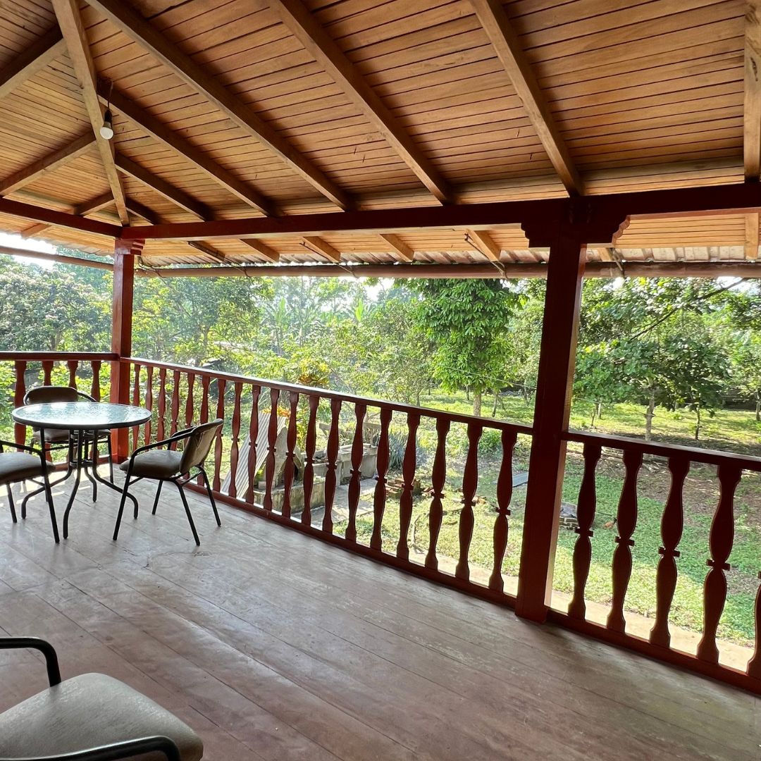 A charming balcony is framed by wooden railings, offering views of surrounding greenery. A round table accompanied by two chairs is positioned on the deck, beneath a sloped wooden roof that contributes to the open atmosphere.