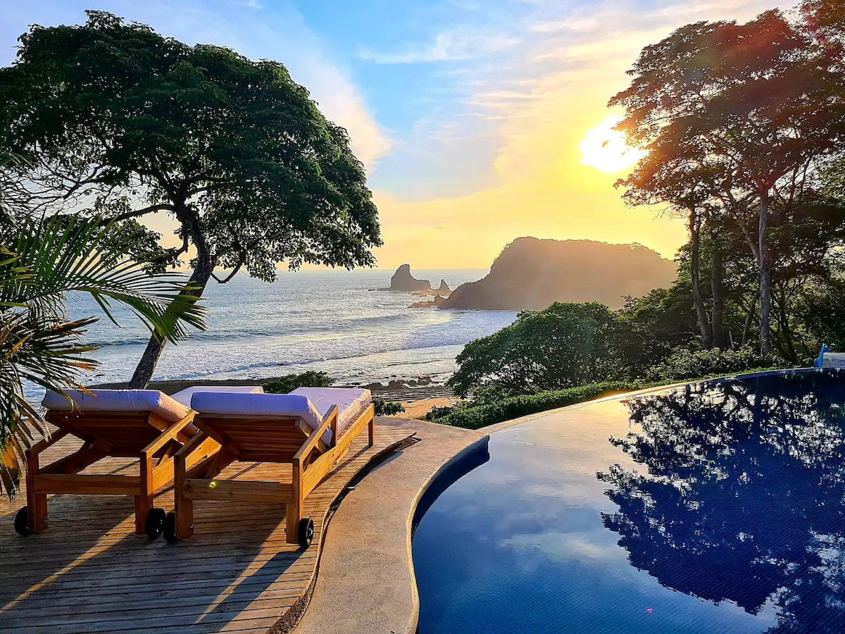 The image captures an infinity pool reflecting the sky, with two wooden loungers positioned at the edge. Lush greenery frames the area, and in the distance, waves gently lap against rocky formations as the sun sets, casting a warm glow over the landscape.