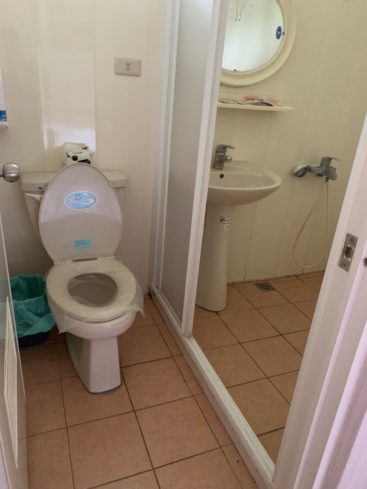 A compact bathroom is visible, featuring a toilet and a sink with a mirror above. The shower area is separated by a glass enclosure. Light beige tiles cover the floor, and a small wastebasket is located next to the toilet.