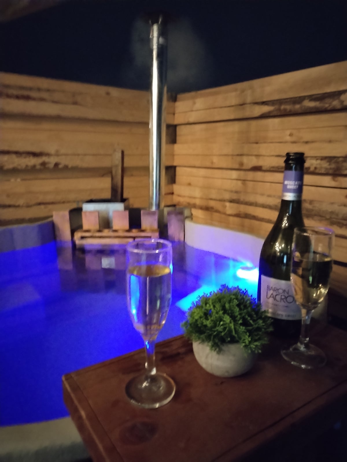 An inviting hot tub is surrounded by wooden paneling, featuring soft blue lighting. Two glasses of sparkling wine rest on a wooden table alongside a small potted plant, creating a serene and relaxing setting.