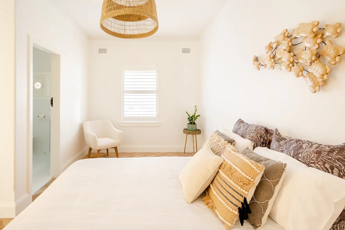 A light and airy bedroom is presented, featuring a spacious bed adorned with decorative pillows. A wicker pendant light hangs above, while a small table with a plant stands beside a chair in the corner. Natural light filters through the window with wooden shutters.