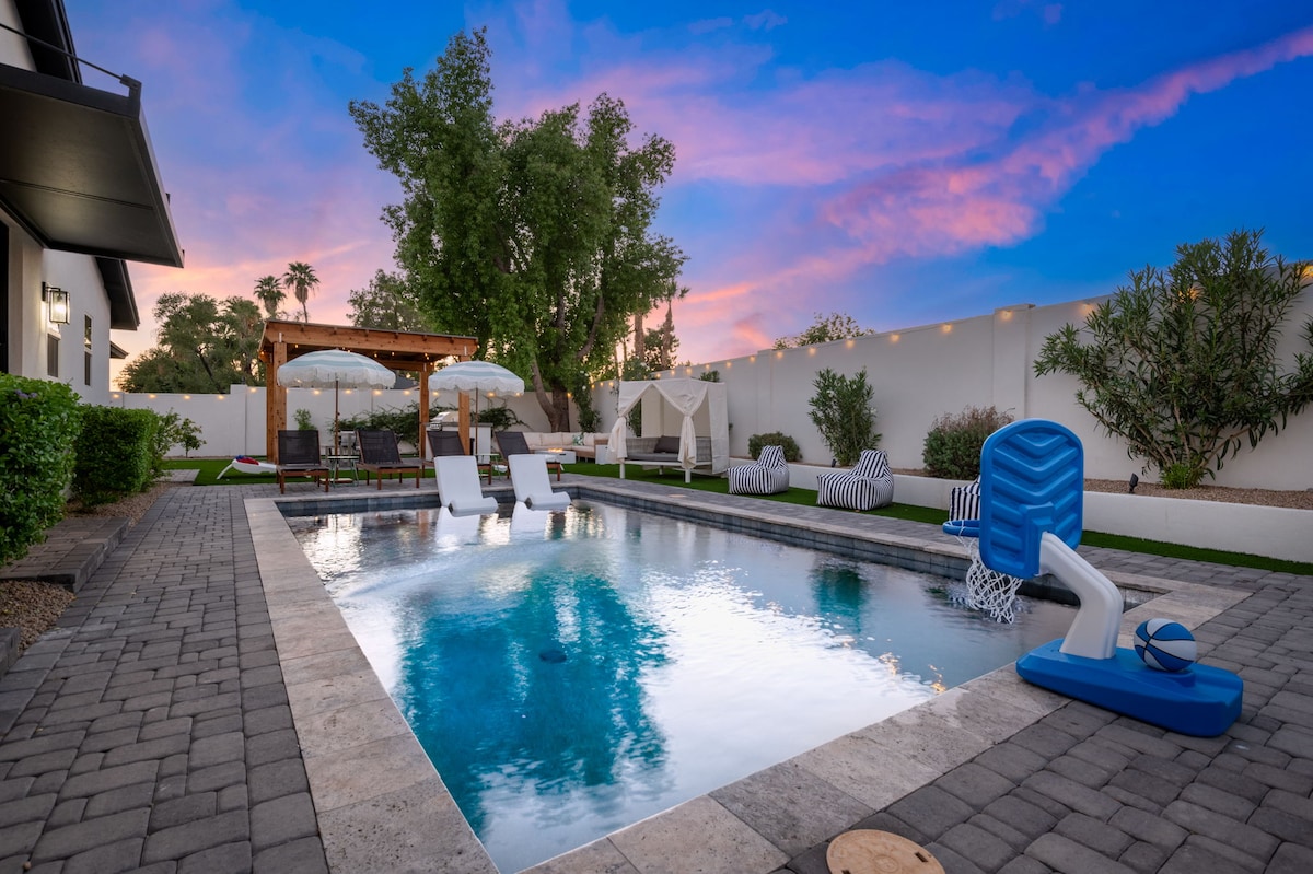 A refreshing pool is surrounded by a spacious patio with loungers and shaded seating areas. A pergola stands at one end, offering a cozy gathering space, while lush trees provide an inviting backdrop. The sky transitions to soft shades of pink and blue at sunset.