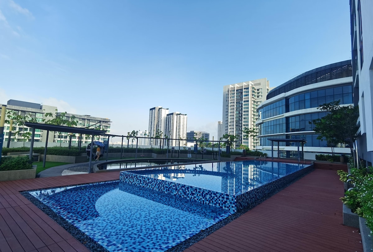 An outdoor pool is set against a backdrop of modern high-rise buildings. The pool features a vibrant mosaic design and is surrounded by wooden decking and greenery, creating a refreshing space for relaxation. The clear blue sky enhances the serene atmosphere.