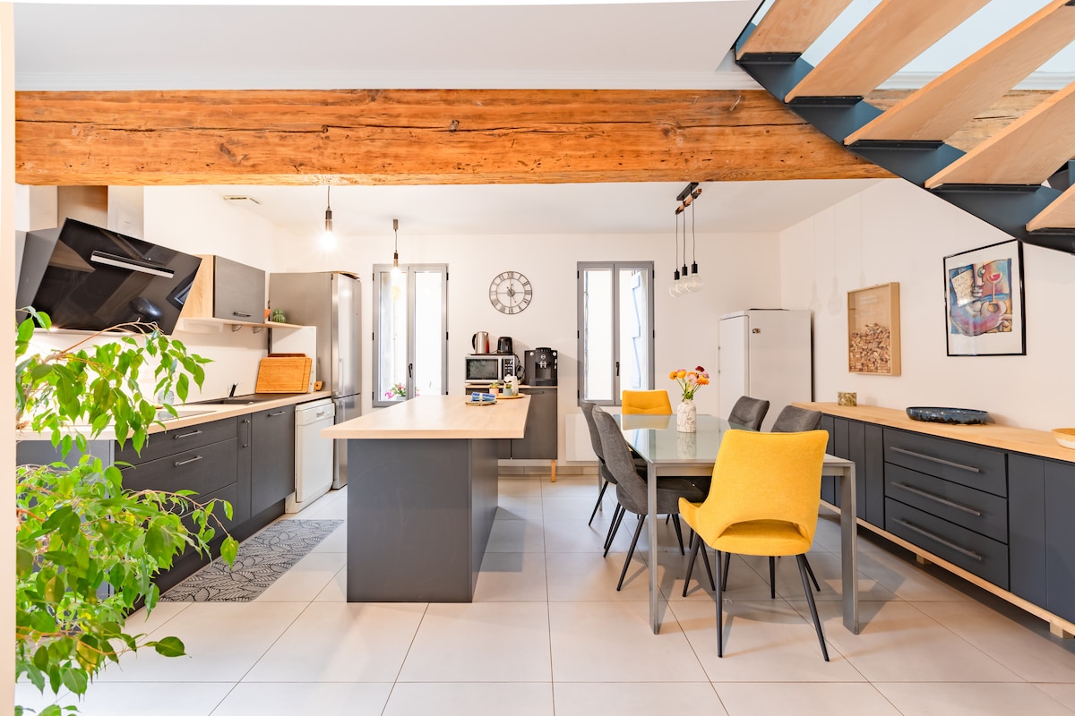 The kitchen and dining area feature modern appliances and a spacious island with seating. Bright yellow chairs surround a wooden dining table. Natural light filters in through the windows, creating a welcoming atmosphere. Wooden beams add character to the room.