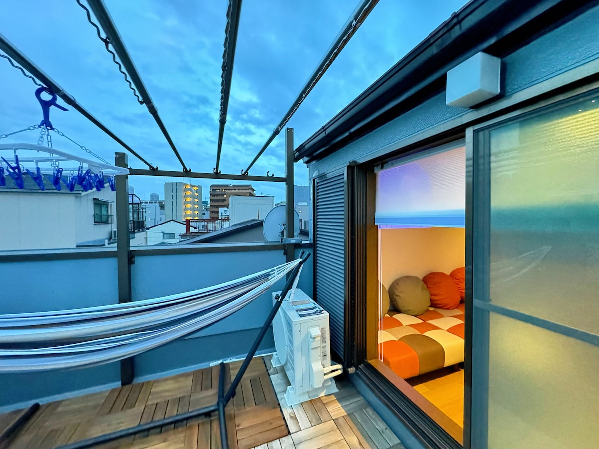 The rooftop terrace features a hammock and a door leading into a cozy indoor space. Soft lighting and a view of the city skyline complement the relaxing atmosphere. Wooden flooring adds warmth, while the open design allows for fresh air and natural light.