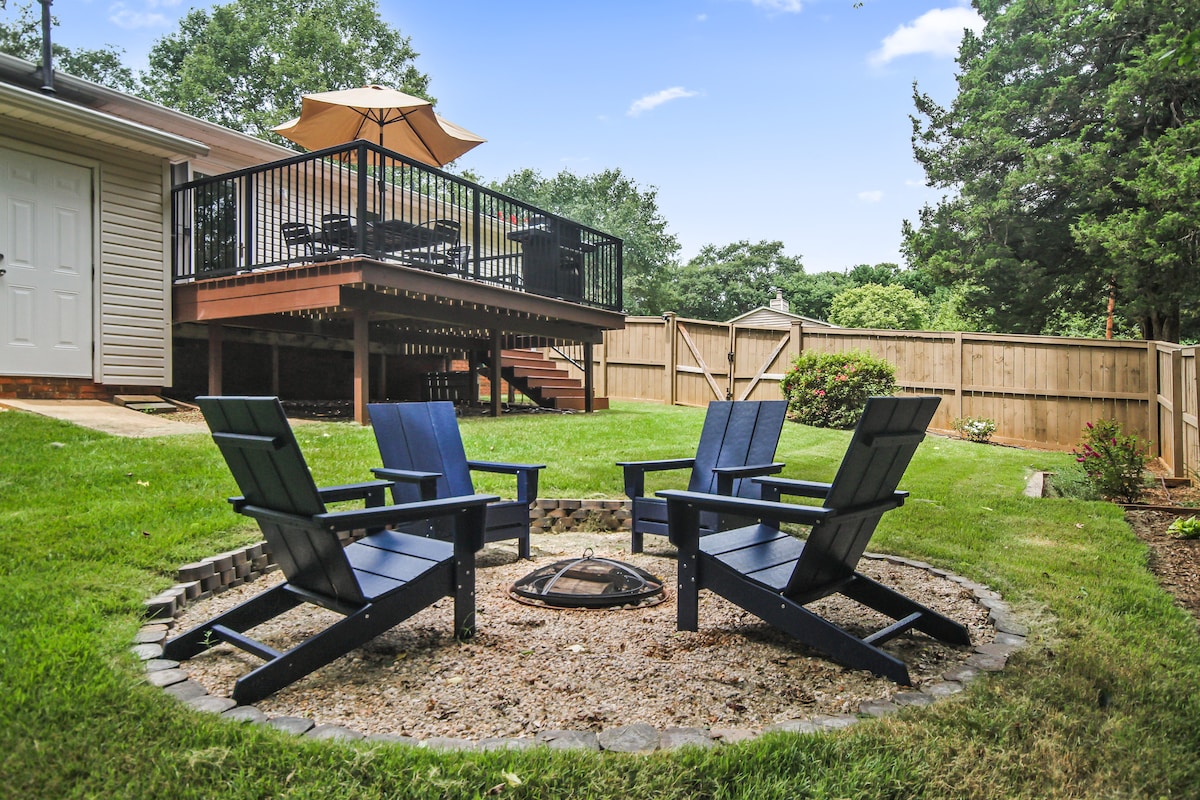 A private backyard offers a cozy firepit area surrounded by four navy blue Adirondack chairs. A wooden deck is visible in the background, featuring an umbrella and outdoor dining space. Lush green grass and flower beds enhance the tranquil outdoor setting.
