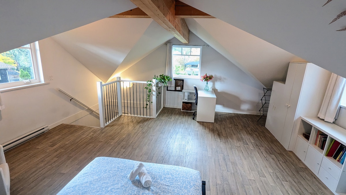 An airy loft space is highlighted by sloped ceilings and wooden beams. Natural light fills the area through large windows, illuminating a workspace with a desk and chair. Greenery adds a touch of life, complemented by shelving and a neatly made bed with fresh towels.