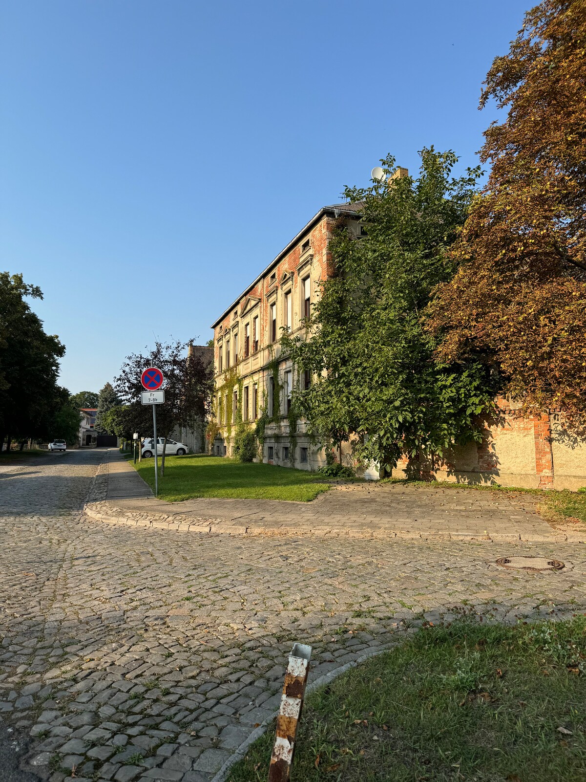 The image features an elegant historic building surrounded by lush green trees. A cobblestone pathway leads to the structure, which displays preserved architectural elements. A street sign is visible in the foreground, indicating a no-parking zone, while a clear blue sky complements the scene.