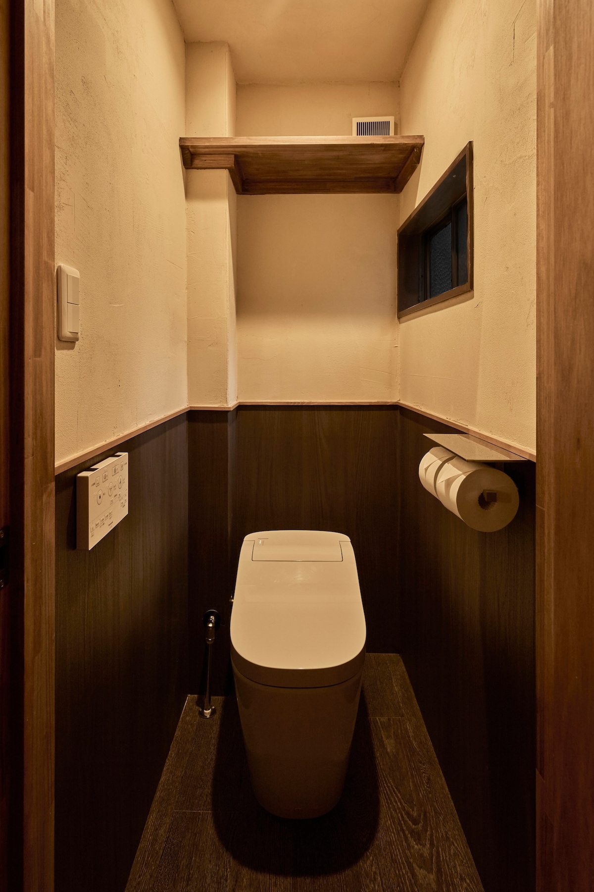 A compact bathroom features modern toilet facilities, paired with a wall-mounted shelf for storage. The warm-toned walls create a calming environment, while a small window offers natural light, enhancing the space. Two rolls of toilet paper are neatly arranged on a holder.