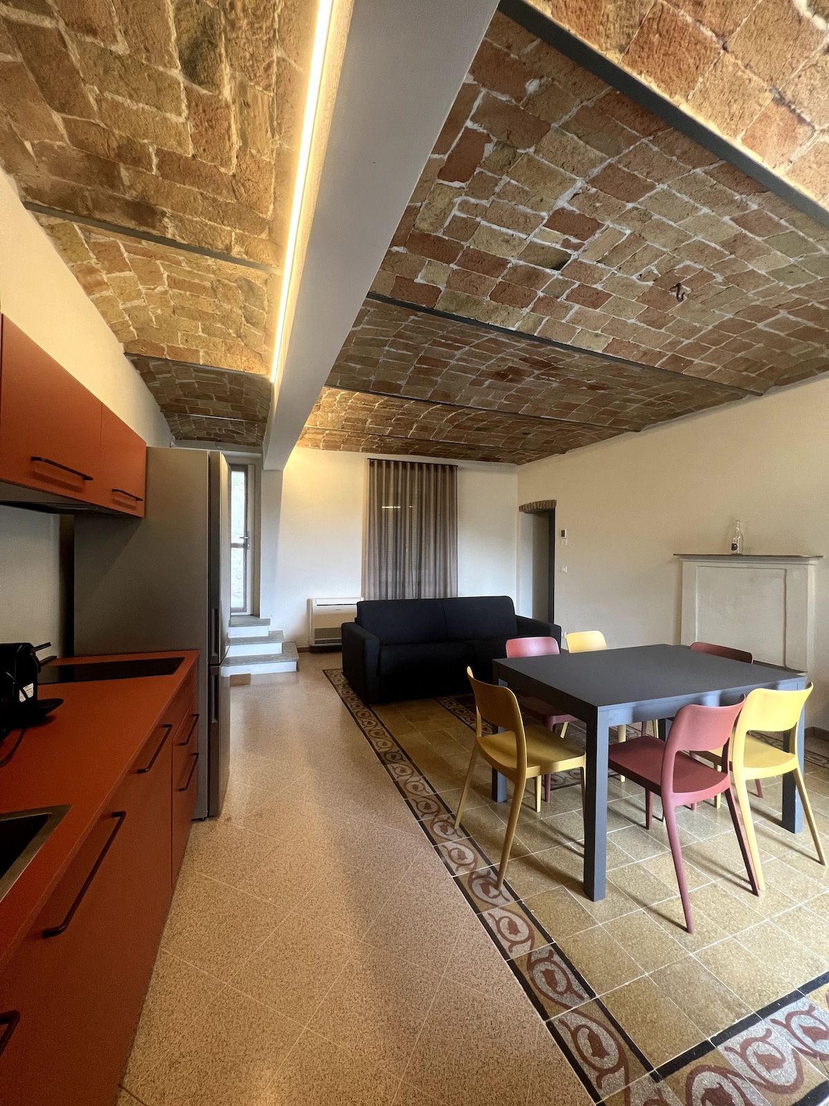 The spacious kitchen features rich red cabinetry and modern appliances. A dining table is surrounded by colorful chairs, complemented by a cozy living area with a dark sofa. The ceiling displays exposed brickwork, enhancing the inviting ambiance of the space.