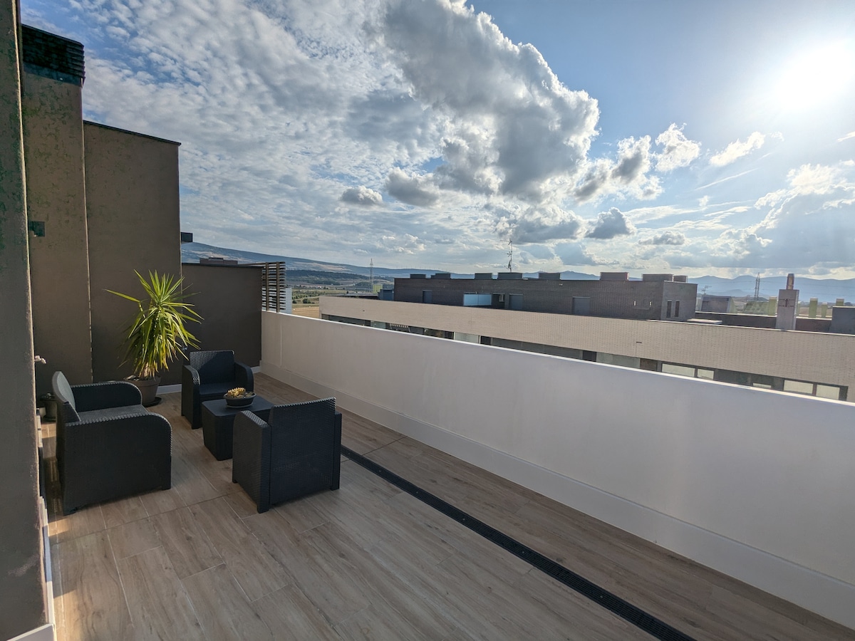 A spacious terrace is presented, featuring comfortable seating arrangements highlighted by a potted plant. Expansive views of the surrounding area are available, with clouds casting shadows across the landscape, and natural light accentuating the sleek flooring.
